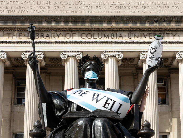 Statue in front of Columbia decorated with a mask, vial, and sign saying 'be well'